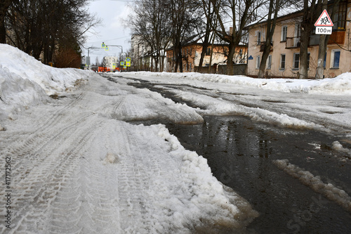 Winter thaw or the beginning of spring. A street in a provincial town. Roadway with large puddles and pits. Dirty ice and snow on the road. Snowdrifts on the sidelines.