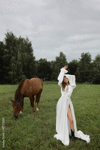 Beautiful young caucasian woman in white gown posing near the brown horse on the green meadow on the summer day © innarevyako