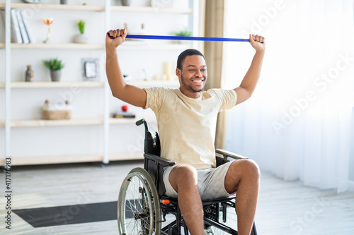 Happy black disabled man in wheelchair doing exercises with rubber band at home