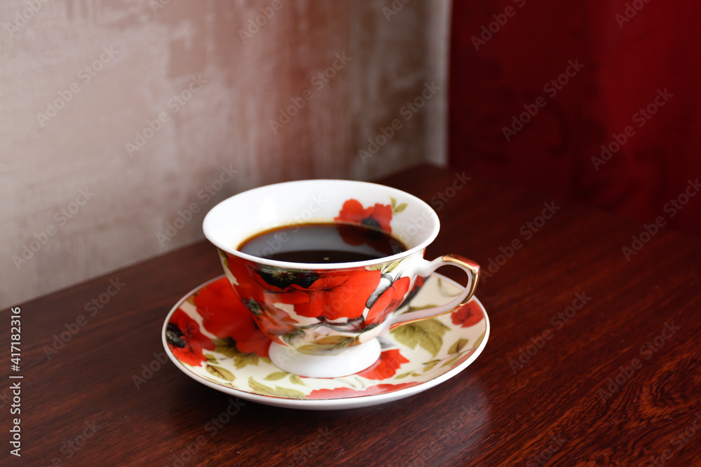 Obraz premium Elegant coffee service with red poppies. A cup of black coffee on the wooden table against the background of red curtain and beige wallpaper.