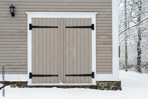 Two grey wooden exterior barn style doors with black wrought iron hinges in an old vintage building. The building has been painted grey with white trim. The foundation has a rock foundation.