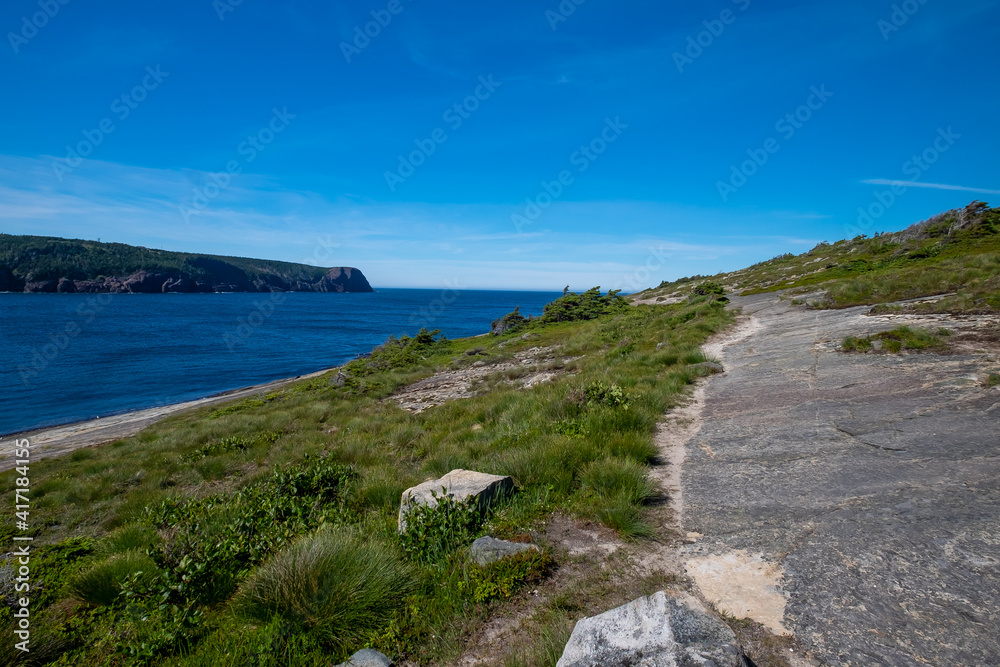 Worn gravel and rocky footpath or hiking trail along a coastline near the edge of blue ocean's water in a cove.  There's a tree covered island in the distance near the horizon with a deep blue sky.
