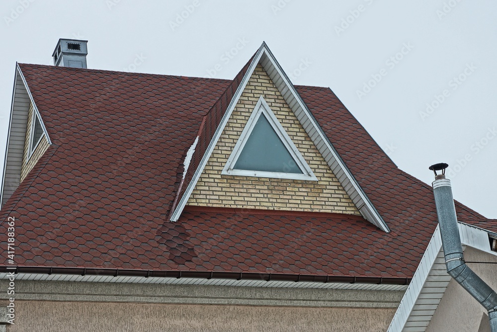 brown brick attic with one triangular window on the tiled roof of a private house against a gray sky