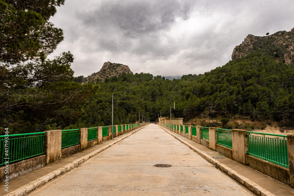 Guadalest reservoir, views of the road and the Mediterranean forest, with some people walking in the background, on a cloudy day.