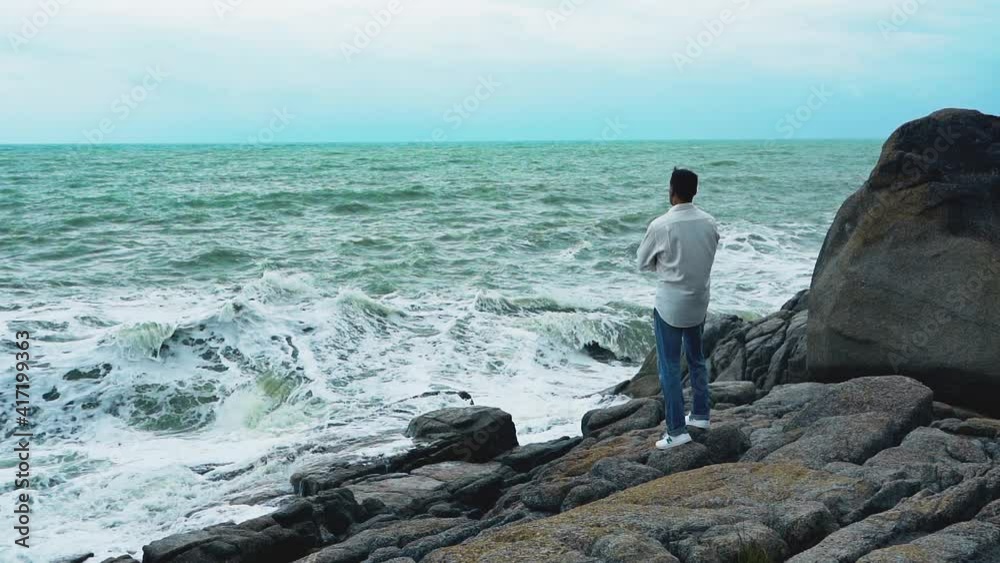 A young man in a white shirt and jeans standing on a rock during a storm and looking at the sea A young man in a white shirt and jeans standing on a rock during a storm and looking at the sea