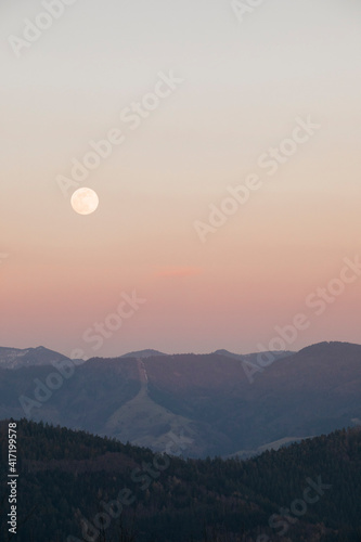 Scenic full moon above mountain peaks with sunset orange sky.