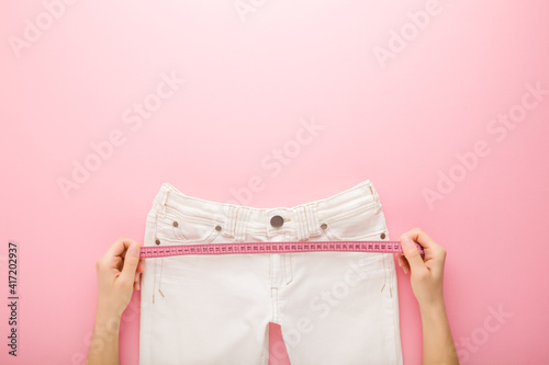 Young adult woman hands holding measure tape and measuring white jeans on light pink table background. Pastel color. Point of view shot. Female weight loss. Empty place for text. Top down view.