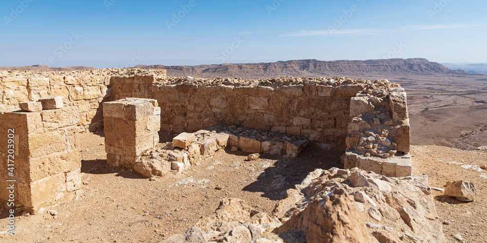interior stone walls and pillars of the ancient Nabatean Makhmal Fortress on the rim of the Maktesh Ramon crater in Israel with Mount Ardon and a hazy blue sky in the background