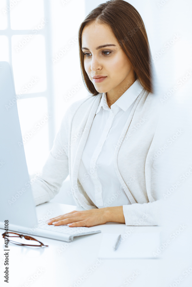 Young cheerful woman sitting at the desk with computer and looking at camera in white colored office. Looks like student girl or business lady. Headshot Young cheerful woman sitting at the desk with computer and looking at camera in white colored office. Looks like student girl or business lady. Headshot