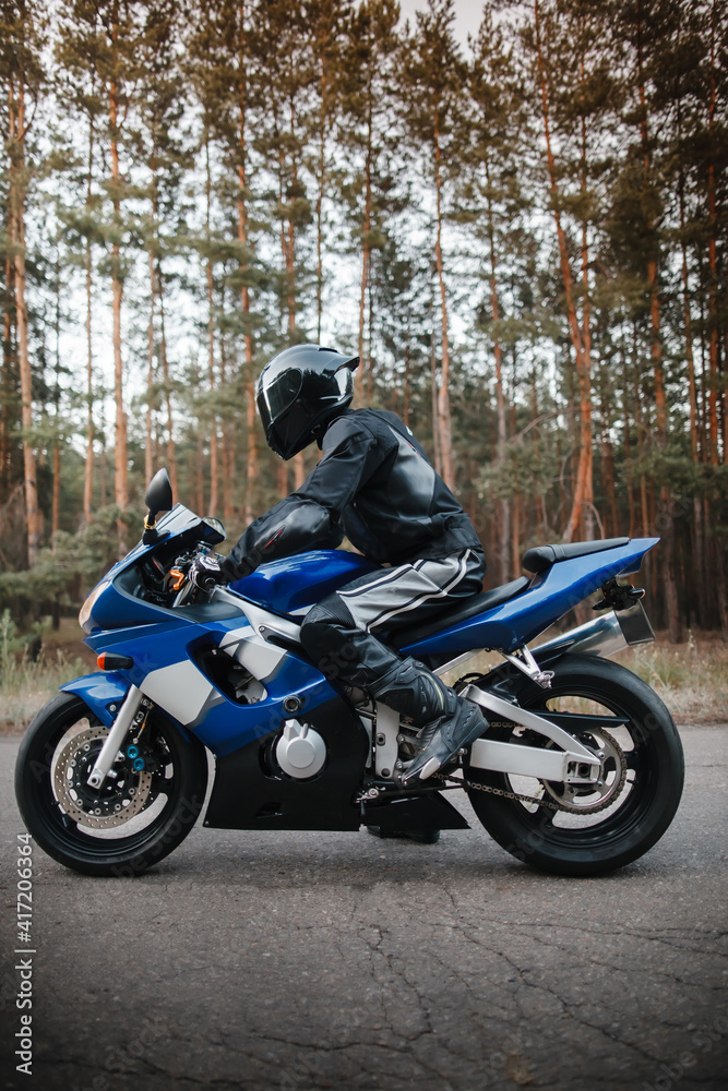 Motorcyclist in leather protective suit and black helmet sits on sports motorcycle. Biker in black rides on the road against the background of the forest. Side view