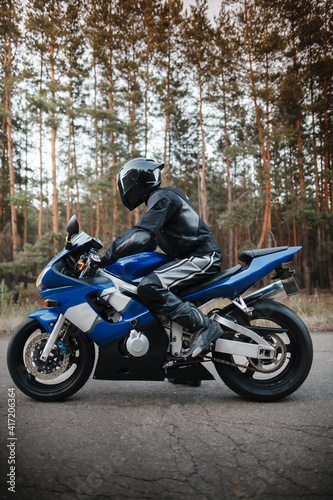 Motorcyclist in leather protective suit and black helmet sits on sports motorcycle. Biker in black rides on the road against the background of the forest. Side view