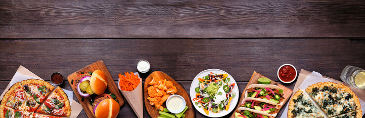 Healthy plant based fast food bottom border. Top view over a dark wood banner background. Table scene with cauliflower crust pizzas, bean burgers, mushroom tacos and vegetarian sides. Copy space.