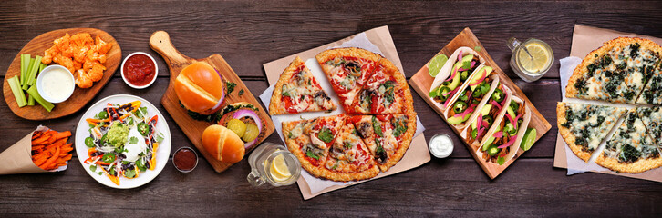 Healthy plant based fast food table scene. Above view on a wood banner background. Cauliflower crust pizzas, bean burgers, mushroom tacos and vegetarian sides.