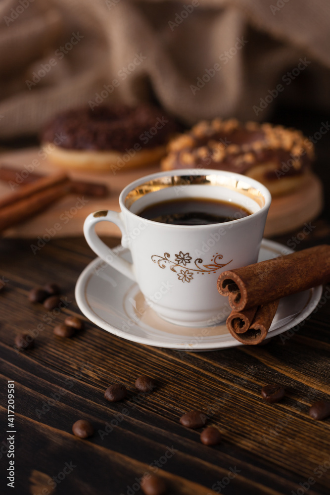 A cup of aromatic black coffee and coffee beans on the table with cinnamon sticks. Morning espresso coffee for breakfast in a beautiful white cup. With a delicious donut in the background. A cup of aromatic black coffee and coffee beans on the table with cinnamon sticks. Morning espresso coffee for breakfast in a beautiful white cup. With a delicious donut in the background.