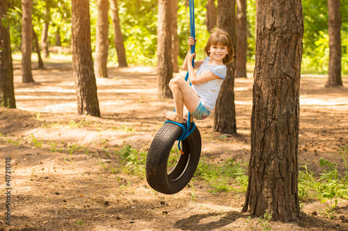 Cute little girl swinging on wheel attached to big tree in autumn forest. 