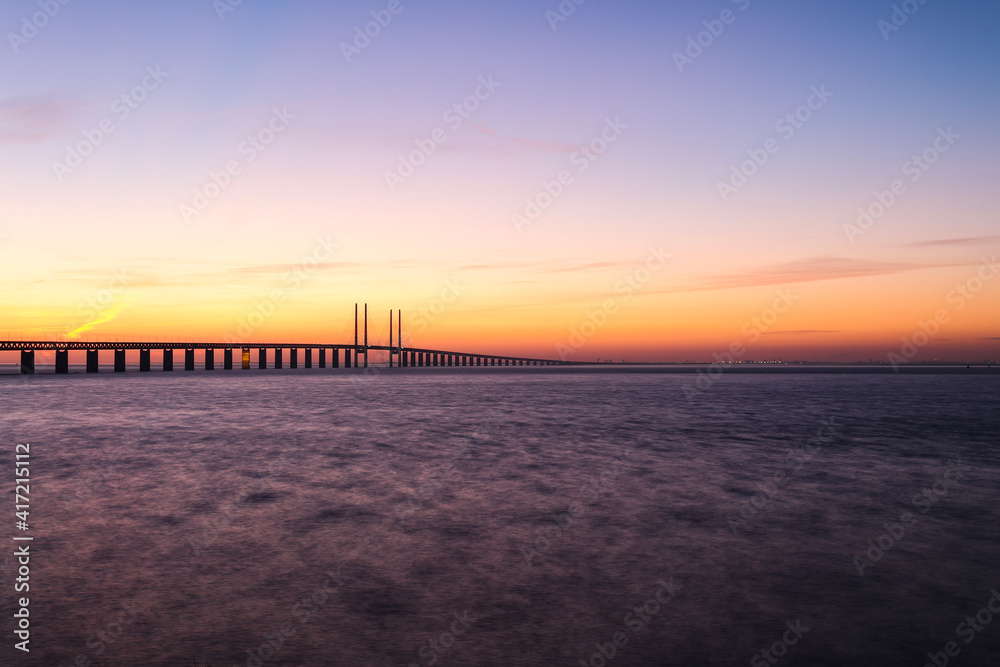 Sunset at the Oresund bridge between Sweden and Denmark at the viewpoint near Limhamn, February 2021. Wide angle, clear sky, bridge stretching from the left to the horizon, long exposure.