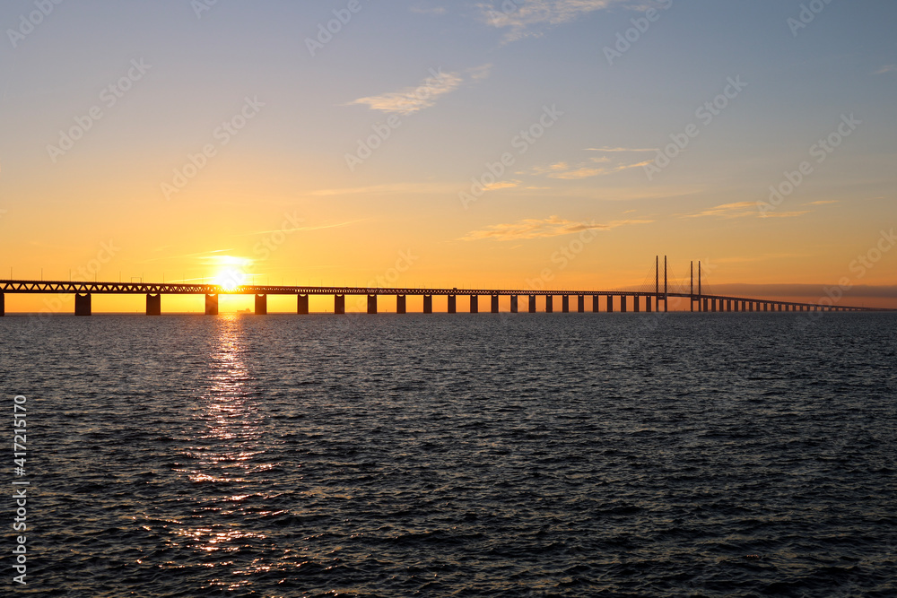 Sunset with the sun over the Oresund bridge at the viewpoint near Limhamn, Sweden, in February 2021. Clear sky, bridge stretching from left to right. Ship visible in the sun rays.