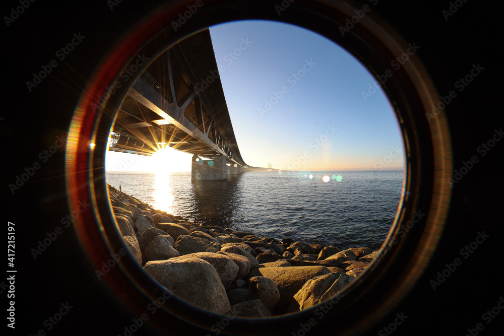 Inside lens view of the Sun under the Oresund bridge Sweden side at the viewpoint near Limhamn, February 2021. Wide angle, clear sky, bridge stretching from the near left to the horizon.