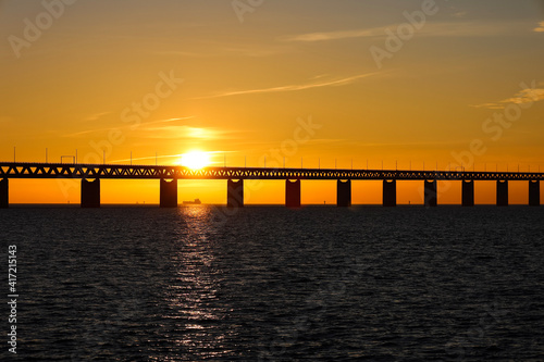 Sunset with the sun over and through a section of the Oresund bridge at the viewpoint near Limhamn  Sweden  in February 2021. Clear sky. Ship visible through the pillars in the evening rays.
