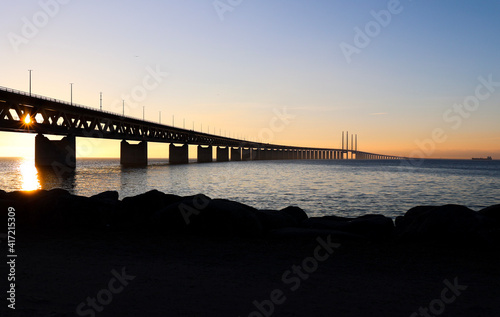 Sun seen through the Oresund bridge at the viewpoint near Limhamn, Sweden, in February 2021. Wide angle, clear sky, bridge stretching from the left to the horizon.