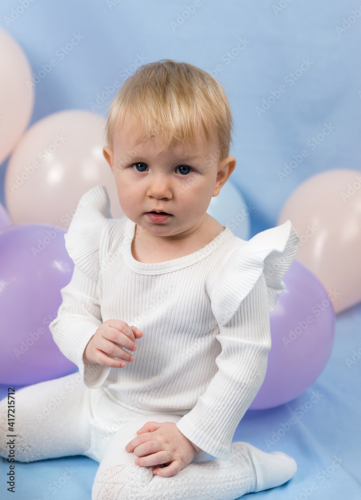 A beautiful serious little girl 1 year old with blue eyes and white turtleneck sits among the balloons and looks at the camera