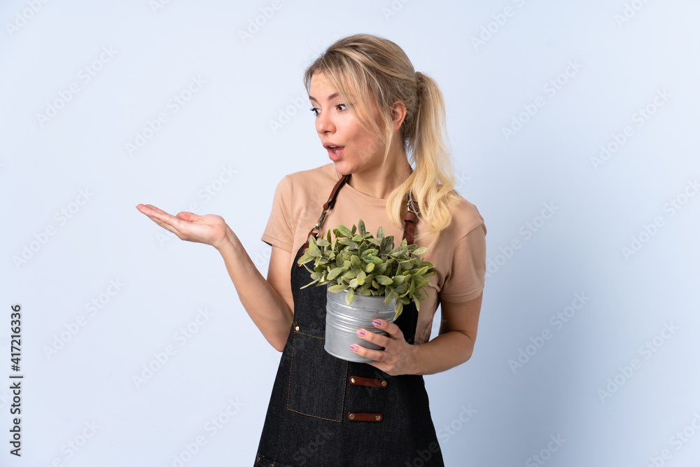 Blonde gardener woman holding a plant over isolated background holding copyspace imaginary on the palm Blonde gardener woman holding a plant over isolated background holding copyspace imaginary on the palm