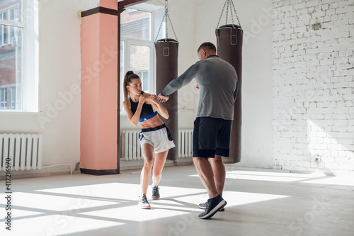 Medium shot of young woman doing boxing workout at the gym and punching her instructor, who is dodging her blows and controlling training process.