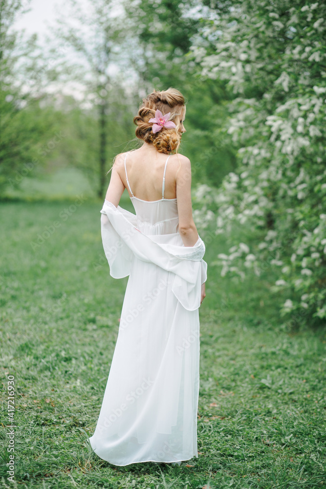 The bride stands with her back to the spring in the park in a white boudoir dress. She is standing by a tree with white flowers