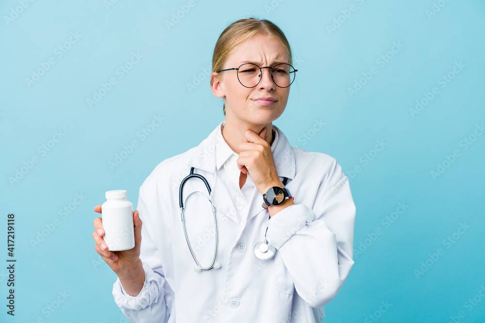 Young russian doctor woman holding pills bottle on blue suffers pain in throat due a virus or infection. Young russian doctor woman holding pills bottle on blue suffers pain in throat due a virus or infection.
