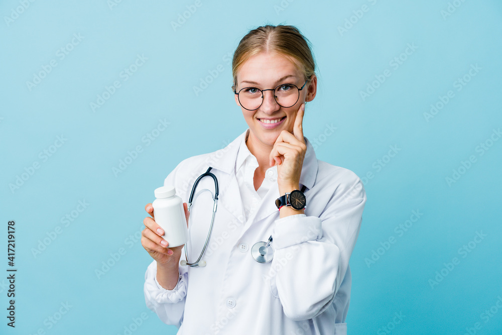 Young russian doctor woman holding pills bottle on blue smiling happy and confident, touching chin with hand.