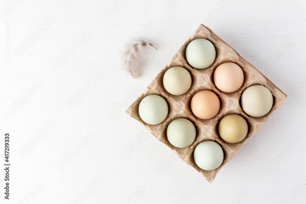 Open carton box with organic eggs and feather on white wood table. Bio food from farm on white wooden background. Copy space, top view. Open carton box with organic eggs and feather on white wood table. Bio food from farm on white wooden background. Copy space, top view.