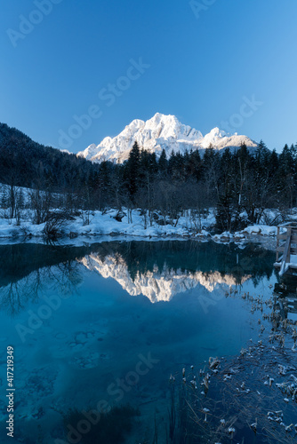 romantic path through snow along a beautiful emerald, clear volcanic lake in winter at a touristic nature reserve in the Alps. Reflection of snowcapped mountains glowing in the rising sun. no people