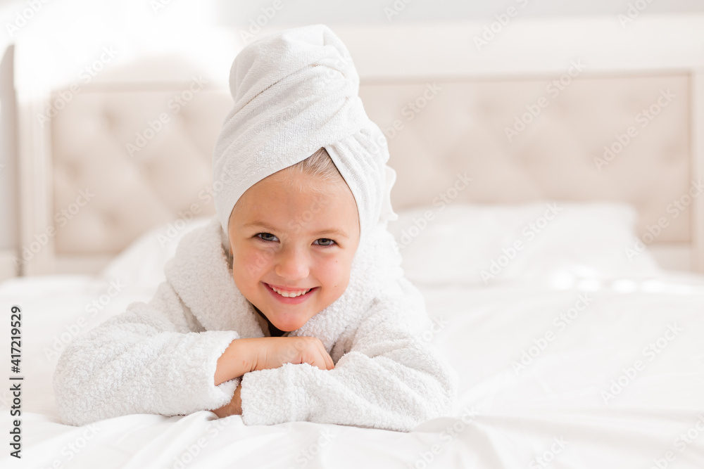 little smiling girl in a white robe with a white towel on her head is lying in bed. portrait of a child little smiling girl in a white robe with a white towel on her head is lying in bed. portrait of a child