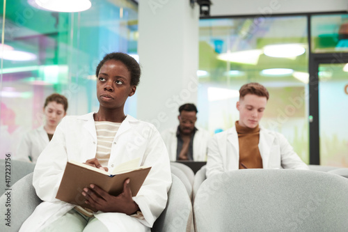 Front view portrait of young African American woman wearing lab coat while sitting in audience and listening to lecture on medicine in college or coworking center, copy space © Seventyfour