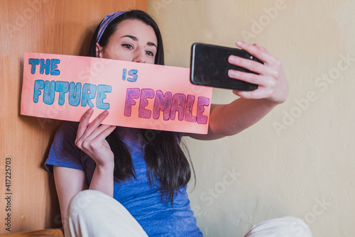 Fotografie Una mujer morena joven vestida de morado se hace una foto con su teléfono móvil mientras sujeta un cartel con un mensaje feminista El futuro es femenino