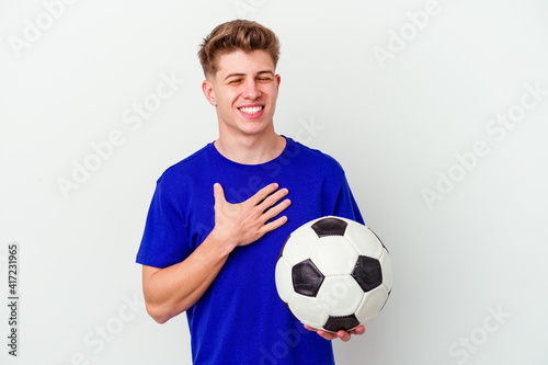 Young caucasian man playing soccer isolated on background laughs out loudly keeping hand on chest.