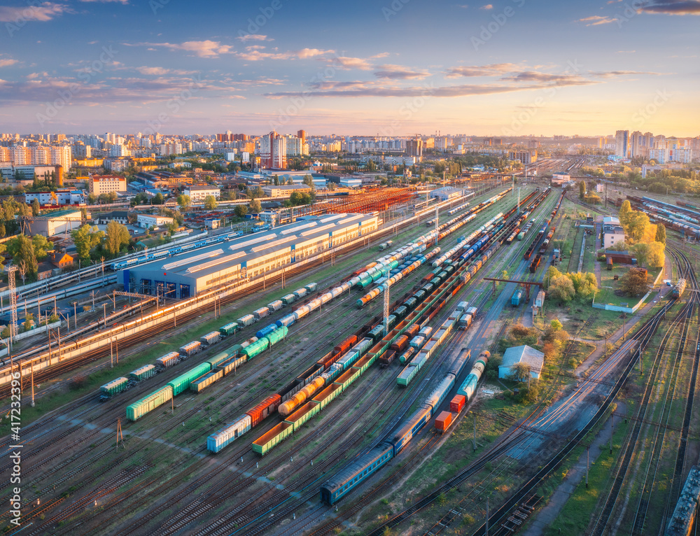 Fototapeta premium Aerial view of freight trains at sunset. Top view of railway station, wagons, railroad. Heavy industry. Industrial landscape with train in depot, city, buildings, colorful sky. Transportation 