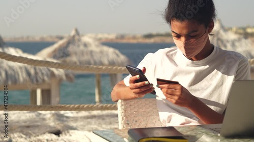 Shop online. Young mixed race woman wearing protective mask making online payment using her mobile phone, looking excited, waiting for her new purchase, sitting on the beach on a sunny day
