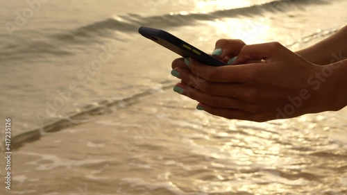 Young woman leaving comments, scrolling news feed, liking posts, using mobile phone with sunrise on the beach in the background. Freelance work, traveler concept
