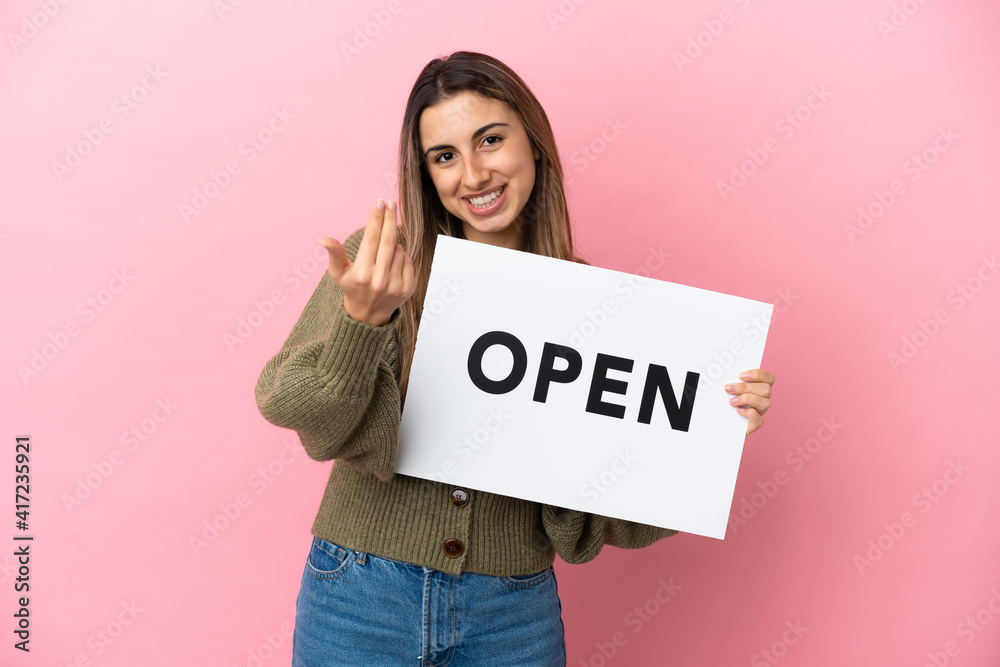 Young caucasian woman isolated on pink background holding a placard with text OPEN and doing coming gesture Young caucasian woman isolated on pink background holding a placard with text OPEN and doing coming gesture