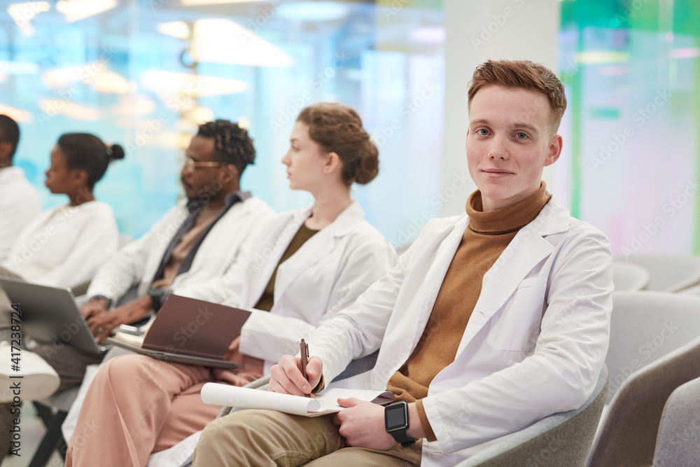 Portrait of young man wearing lab coat and smiling at camera while sitting in row with multi-ethnic group of people in audience at medical seminar, copy space Portrait of young man wearing lab coat and smiling at camera while sitting in row with multi-ethnic group of people in audience at medical seminar, copy space