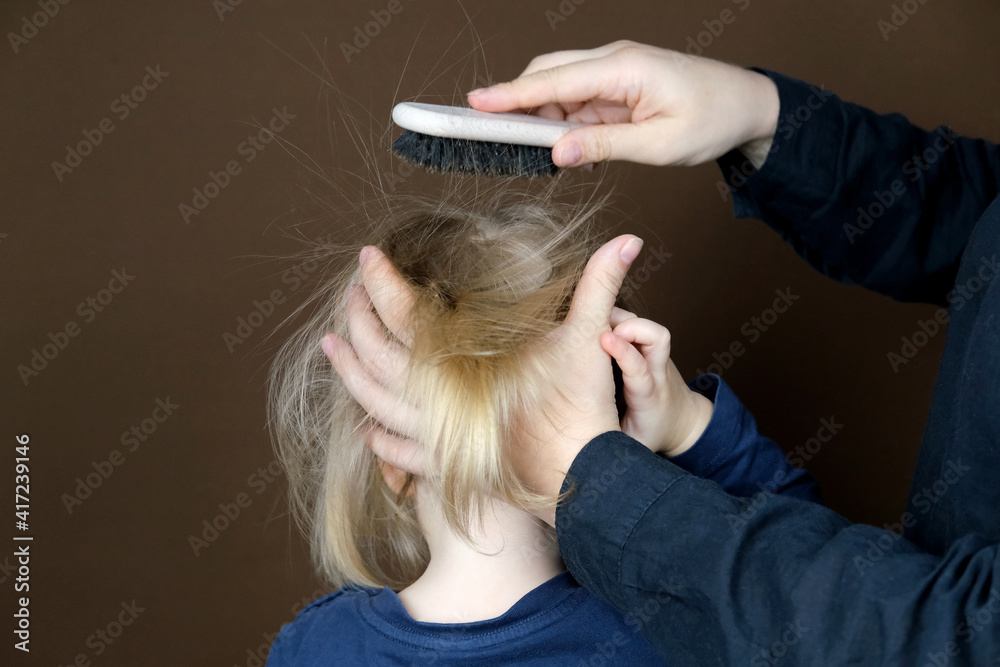 Fototapeta premium mom combing her daughter's hair, little blonde girl with a wooden brush with natural bristles, child and woman on a dark brown background, concept of hygiene, hair care, childhood, motherhood