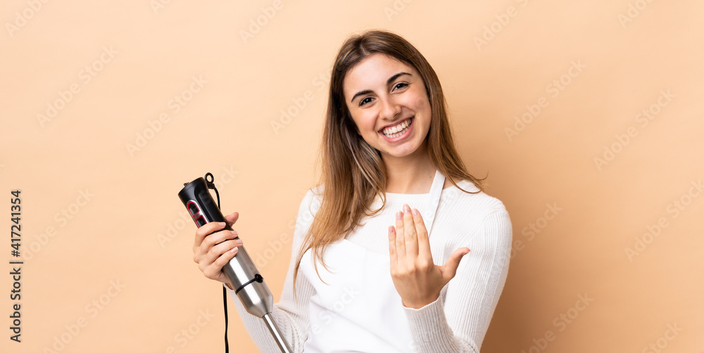 Woman using hand blender over isolated background inviting to come with hand. Happy that you came Woman using hand blender over isolated background inviting to come with hand. Happy that you came