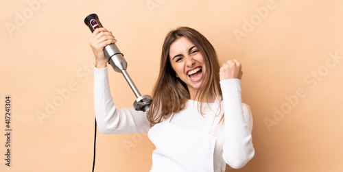 Woman using hand blender over isolated background celebrating a victory