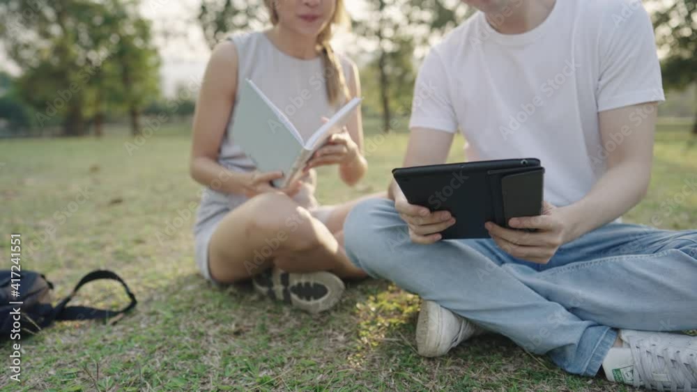 Young colleague working together on the outdoor park, sit down the ground with green grass, discuss brainstorm helping hand on the project, using tablet doing research, classmate reviewing books Young colleague working together on the outdoor park, sit down the ground with green grass, discuss brainstorm helping hand on the project, using tablet doing research, classmate reviewing books