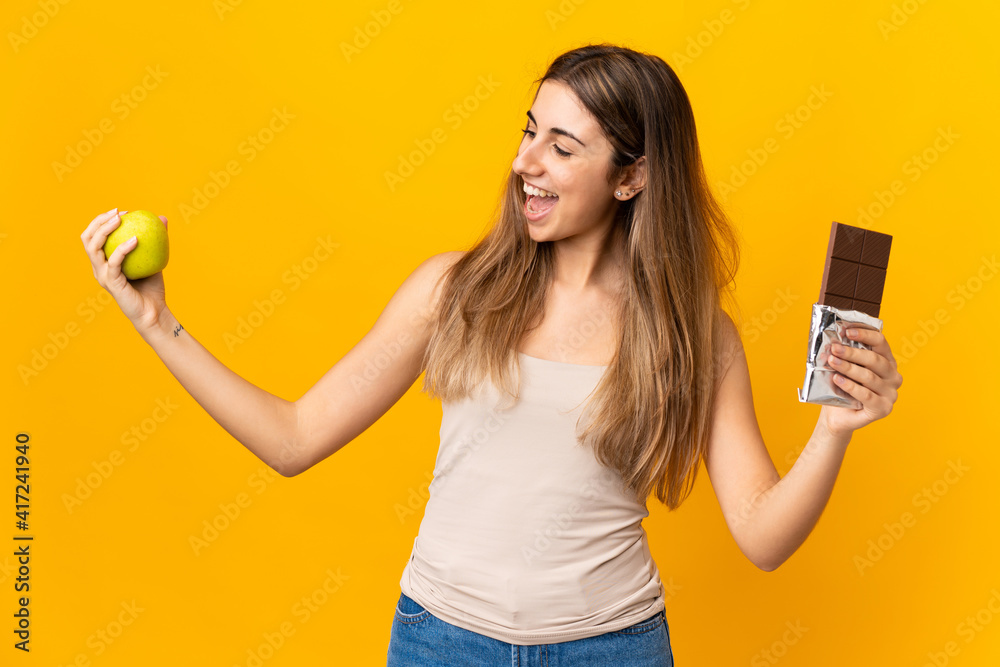 Young woman over isolated yellow background taking a chocolate tablet in one hand and an apple in the other Young woman over isolated yellow background taking a chocolate tablet in one hand and an apple in the other