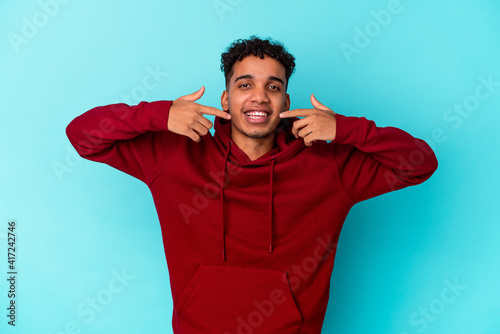 Young african american curly man isolated on blue smiles, pointing fingers at mouth.