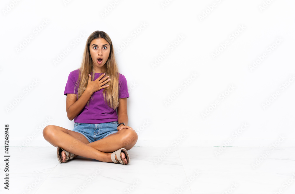 Young hispanic woman sitting on the floor surprised and shocked while looking right