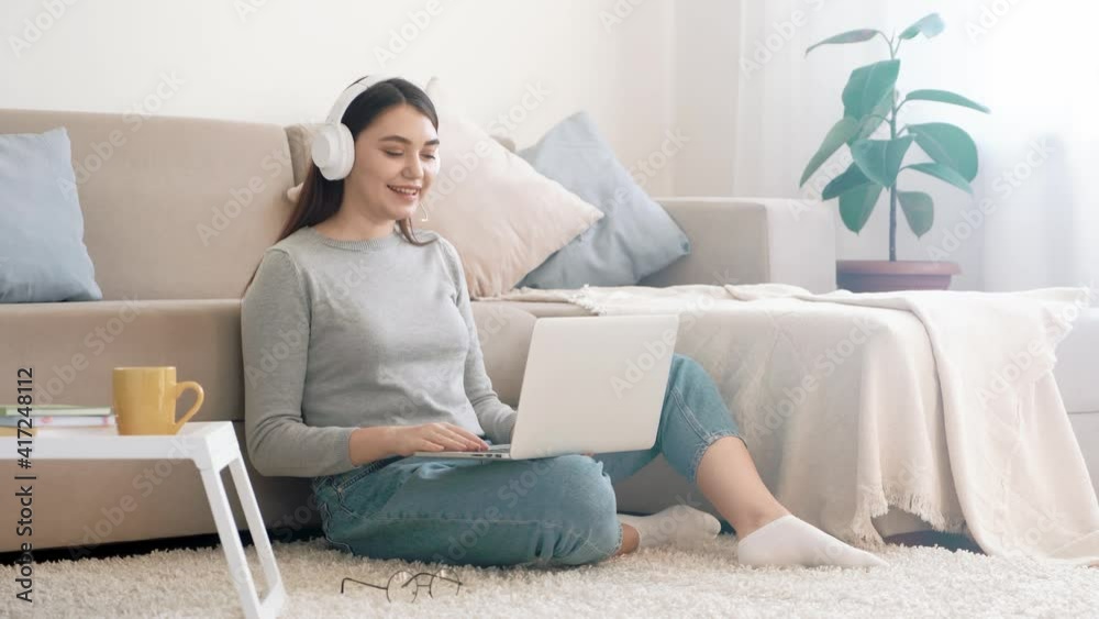 Smiling young woman in wireless headphones sit on the floor and communicating with friends via videocall on laptop Smiling young woman in wireless headphones sit on the floor and communicating with friends via videocall on laptop