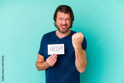 Middle age dutch man holding a help placard isolated on blue background showing fist to camera, aggressive facial expression.